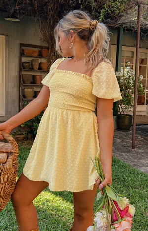 Blonde haired model is pictured wearing a yellow mini dress. The dress features bell top sleeves and a ruched bust. The fabric is multi textured across the whole dress.